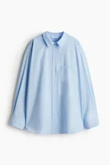 Camisa celeste de algodón suave, de corte holgado, con cuello clásico, botones frontales, canesú en la espalda, hombros caídos, bolsillo de parche en el pecho, mangas largas con puños abotonados y bajo redondeado.
