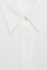 Camisa blanca de cuello solapa y manga corta, con bolsillo delantero y detalle de vainicas a tono. Cierre frontal con botones ocultos por solapa.