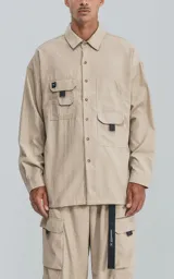 Camisa de manga larga color beige, estilo técnico con calce oversize. Presenta cuello camisero, cierre frontal de botones y múltiples bolsillos de parche con solapa en el frente, algunos con tiradores negros.