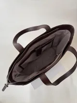 Cartera tipo tote color beige, hecha en cuero graneado con textura uniforme y suave al tacto. Tiene doble asa de hombro, un bolsillo frontal con broche imantado, un bolsillo trasero con cierre metálico y cierre principal también metálico. El interior está forrado y cuenta con tres bolsillos: uno con cierre, uno tipo plaqué para celular y un bolsillo grande tipo sobre con solapa y botón.