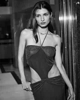 Vestido largo negro, ajustado al cuerpo, con escote strapless y tirantes finos cruzados en el cuello. Presenta recortes laterales pronunciados en la cintura y un nudo fruncido en la parte inferior frontal.