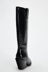 Bota alta negra de cuero sintético, estilo cowboy, con punta fina y tacón geométrico.