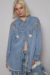 Camisa de jean celeste de corte amplio, con botones frontales y detalles de costuras desgastadas. Presenta apliques de flores tejidas en el pecho y terminación desflecada.