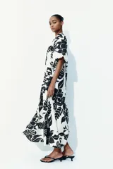 Vestido largo de corte holgado con estampado floral en blanco y negro. Tiene escote cuadrado con cordón ajustable y mangas cortas abullonadas con elástico en los puños. Presenta costura fruncida bajo la cadera.