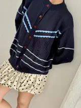 Saco de punto calado color beige claro, con cuello redondo y cierre frontal de botones azules. Presenta detalles decorativos en crochet verde y negro en el frente y puños.