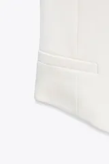 Top tipo chaleco blanco de tejido de punto elástico, con escote asimétrico, manga sisa y cierre frontal con botones dorados.