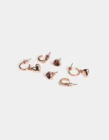 Set de tres pares de aros bañados en oro rosa: un par de aros tipo cuff con strass, un par de aros tipo botón con forma de corazón y cristal color borgoña, y un par de aros colgantes tipo argolla con dije de corazón liso.