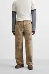 Pantalón de jean color beige con estampado abstracto floral en tonos marrones, de corte amplio, con bolsillos delanteros y traseros, y detalle de tira multifunción en la pierna. Cierre frontal con cremallera y botón.