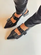 Zapato negro de cuero sintético, con punta fina, escote asimétrico y dos tiras con hebillas metálicas.