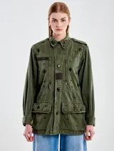 Chaqueta militar verde oliva de algodón con cuello camisero, cierre frontal con botones y cuatro bolsillos delanteros con solapa. Decorada con apliques metálicos y parches de tela.