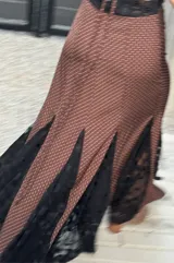 Vestido largo de seda color marrón con estampado de pequeños puntos blancos. Posee una silueta fluida con torso entallado, escote profundo y espalda descubierta con tiras cruzadas. El diseño se destaca por sus inserciones de encaje negro en el ruedo que aportan volumen y movimiento.