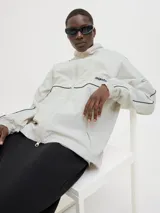 Campera deportiva color crema, de corte recto y cuello alto. Presenta cierre frontal, bolsillos laterales y mangas largas con puños elásticos. Detalle de líneas negras en hombros y mangas, y logo estampado en el pecho.