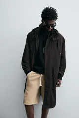 Trench relaxed fit negro confeccionado en tejido técnico ligeramente acolchado, con cuello subido y capucha desmontable. Tiene manga larga, bolsillos de vivo en pecho y de solapa en cadera, y abertura en el centro de la espalda. El cierre frontal es con cremallera oculta por solapa con botones a presión.