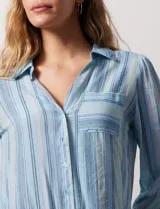 Camisa de gasa celeste con estampado de rayas verticales en tonos azules, rosas y verdes. Tiene cuello con solapa, escote en V, abertura central con botones, bolsillo aplicado en el pecho y terminación anudada en la parte delantera.