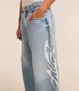 Pantalón de jeans celeste de corte amplio, con cintura con trabillas, cierre tradicional y bolsillos. Presenta estampa lateral estilo maorí en color blanco.