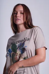 Remera color beige de algodón con cuello redondo y mangas cortas. Estampado de una flor de magnolia y hojas en tonos verdes, azules y rosados. Texto 'Magnolia' en color negro en la parte superior del estampado.