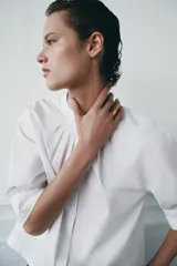 Camisa blanca de algodón con cuello redondo y manga corta abullonada. Presenta un canesú con pliegues verticales en la parte delantera y cierre frontal con botones ocultos. El bajo es redondeado.