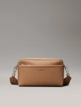 Bandolera Calvin Klein color tostado, con correa ancha de tela con logo de la marca y correa fina de cuero sintético.