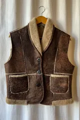 Chaleco marrón de cuero genuino con interior de piel sintética color beige, cierre frontal con botones y dos bolsillos delanteros.