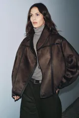 Cazadora de estilo aviador con cuello subido, manga larga y cierre frontal asimétrico con cremallera. Presenta un diseño de doble faz con exterior de efecto ante y forro interior de borreguillo a tono, con detalles de ribetes en cuero sintético.