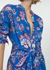 Vestido camisero azul con estampado floral en tonos rosa, naranja y marrón. Tiene cuello mao, cierre frontal con botones, mangas largas con puños abotonados y lazo decorativo en la cintura.