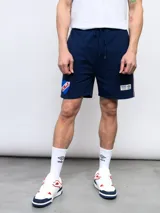 Bermuda deportiva azul marino con cordón ajustable en la cintura y bolsillos laterales. Presenta el escudo del Club Nacional de Football bordado en la pierna izquierda y un logo de Umbro en la pierna derecha. Tiene una franja blanca y roja en los laterales.