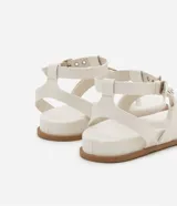 Sandalia plana de cuero sintético color blanco, con tiras superpuestas, detalle de aro metálico y cierre ajustable con hebilla.