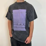 Remera negra con efecto lavado y estampado rectangular con imagen de surfistas en la playa y texto "Visions" en color lila. Corte oversize.