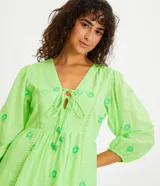 Vestido corto color verde lima con estampado de flores en verde claro, escote en V con lazo, mangas 3/4 con puños elásticos y falda con volantes.