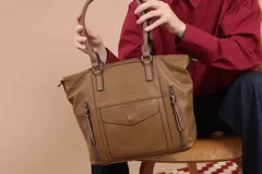 Cartera bandolera de símil cuero color marrón claro, con doble asa rígida y correa regulable. Presenta dos bolsillos frontales con cierre vertical y un bolsillo frontal con solapa y botón. Cuenta con dos divisiones internas.