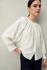 Camisa color crema de lino con cuello redondo, canesú fruncido, mangas largas amplias con puños elastizados con volado y cierre frontal con botones.