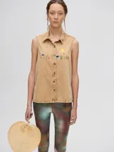 Camisa sin mangas de denim liviano color camel, con cuello camisero y botones frontales. Presenta bordados temáticos marinos en el pecho, incluyendo un sol, olas, una gaviota y vida marina.