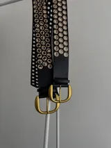 Cinturón negro de cuero ecológico con hebilla dorada y múltiples ojales dorados.