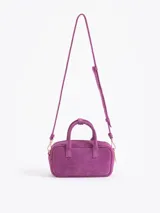 Cartera tipo baulito de cuero descarne vacuno en color fucsia, con diseño compacto y estructura rígida. Presenta doble asa de mano, logo de la marca grabado en relieve en el frente y un charm metálico en forma de vaquita de San Antonio colgando de una de las asas.