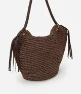 Bolso de playa tipo canasta, tejido en fibra de papel color marrón oscuro, con forma redondeada y abertura superior curva. Cuenta con asas trenzadas de material sintético marrón oscuro y detalles de flecos colgantes a los lados.