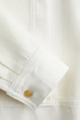 Blusa blanca de corte holgado en sarga de algodón con costuras visibles adelante y atrás. Modelo con cuello redondo, hombros caídos, tapeta con botones ocultos y mangas largas con puños abotonados. Bajo levemente redondeado con abertura pronunciada en los laterales. Espalda algo más larga.