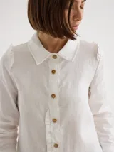 Camisa blanca de lino con cuello clásico, abotonada al frente con botones de madera, mangas largas con volados en los hombros y corte femenino.