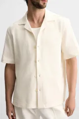 Camisa color crudo de corte regular fit, confeccionada en tejido con textura. Tiene cuello bowling, manga corta, bajo con aberturas laterales y cierre frontal con botones.