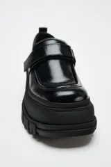 Zapato tipo mocasín negro con plataforma y suela track. Presenta tira ajustable adherente y tirador trasero.