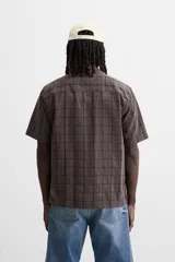 Camisa de hombre color gris con diseño a cuadros, corte relaxed fit, cuello bowling, manga corta y cierre frontal con botones.