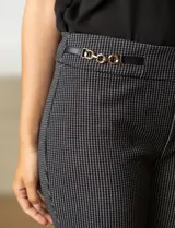 Pantalón largo de vestir negro con finas rayas verticales blancas, corte flared de tiro alto y aplique de cadena dorada en la cintura.