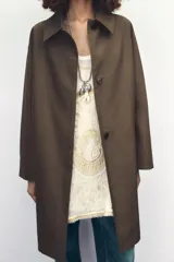 Trench color marrón con cuello solapa y manga tres cuartos. Tiene bolsillos delanteros y cierre frontal con cremallera y botones ocultos por solapa.