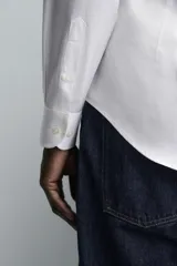 Camisa blanca de algodón, manga larga, con cuello abotonado (button-down) y cierre frontal de botones.