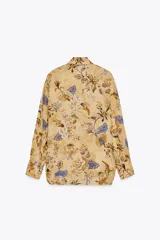 Camisa de ramio color beige con estampado floral en tonos marrones y azules, cuello solapa, manga larga con puños abotonados, bajo asimétrico con aberturas laterales y cierre frontal con botones.