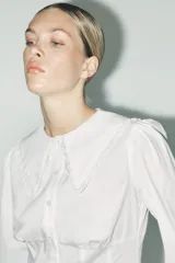 Camisa blanca de popelina de algodón con cuello bobo y mangas largas con elástico en los puños. Detalle de puntilla a tono en cuello y ruedo. Cierre frontal con botones.