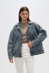 Campera de jean celeste con cuello clásico, solapas, cierre frontal con botones metálicos y mangas largas con puños abotonados. Presenta detalles de costuras diagonales y apliques de tachas metálicas.