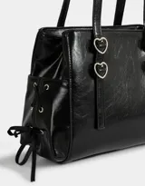 Cartera tipo city de cuerina color negro con efecto charol. Tiene manijas superiores con apliques metálicos en forma de corazón, cierre superior y bolsillos laterales con trenzado decorativo.