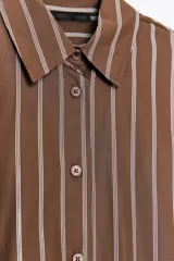 Camisa oversize marrón con rayas verticales blancas, cuello solapa, manga larga con puños y hombros caídos. Tiene un bolsillo de plastrón en el delantero, pinzas en la espalda y cierre frontal con botones.