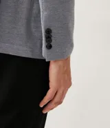 Blazer masculino color gris claro, de tejido liso con textura casual, con solapas, bolsillos amplios de parche en la parte delantera y un bolsillo superior. Cierre frontal de un solo botón.