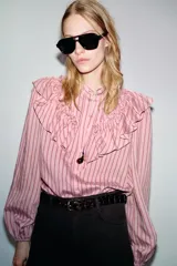 Camisa de manga larga color rosa claro con finas rayas verticales en tono rosado más oscuro. Presenta cuello mao y un gran volante fruncido que recorre el frente, desde los hombros hasta el centro. Los puños son elásticos.